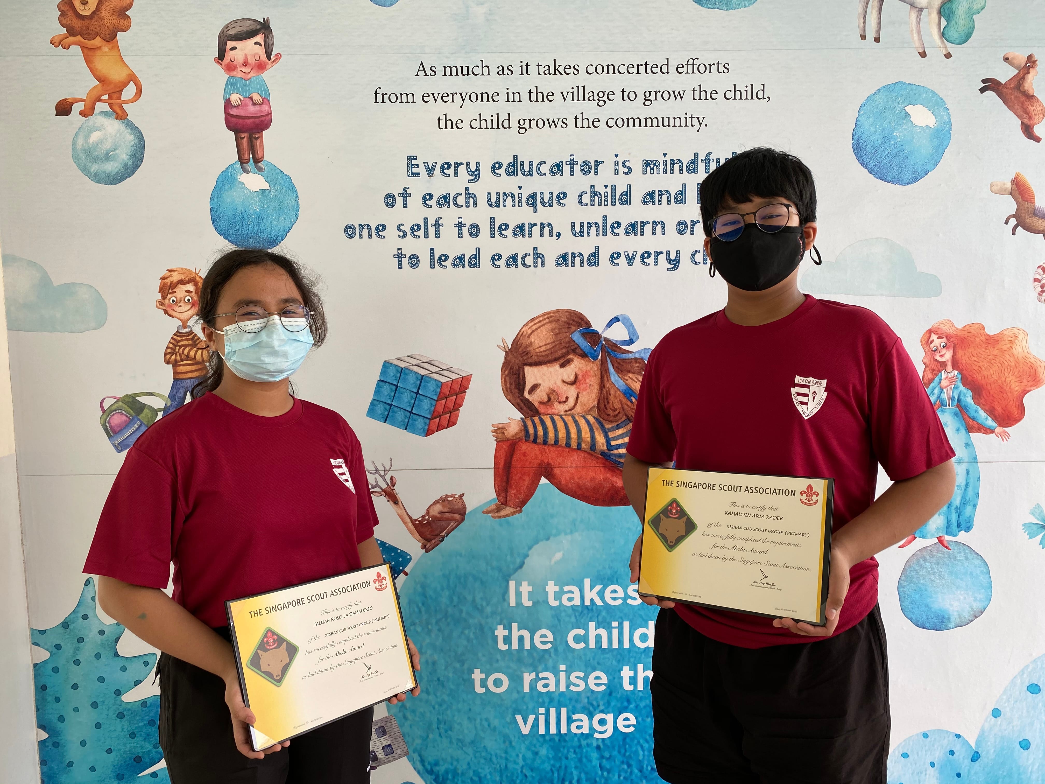 Two masked students in red shirts hold Singapore Scout Association certificates in front of a colorful mural.